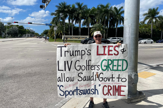 A person holds a sign at the corner of a sidewalk that says &quot;Trump's lies + LIV golfers greed allow Saudi Govt. to 'Sportswash' crime.&quot;