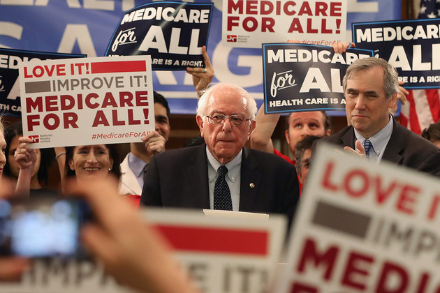 Bernie Sanders stands while people around him hold signs that say Medicare For All. 