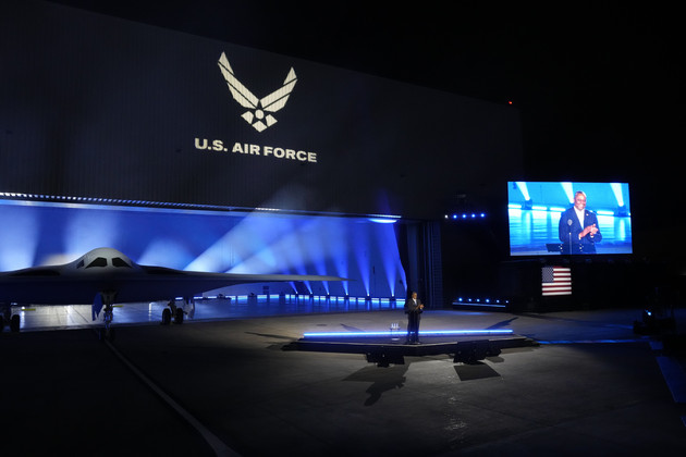 A B-21 Raider is pictured on display as Defense Secretary Lloyd Austin speaks at a microphone with his image projected onto a screen.