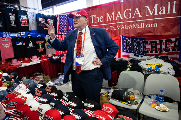 Ronald Solomon speaking from his MAGA merchandise stand, surrounded by hats, shirts, and flags.