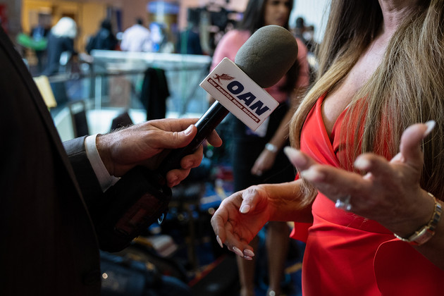 A close up of Kimberly Guilfoyle's hands gesturing while speaking into an One America News-branded microphone.